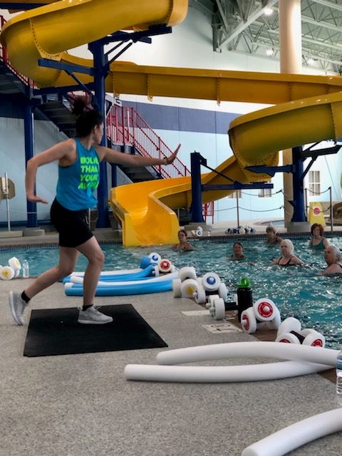 Image depict instructor on pool deck with class participants in the water using equipment to workou