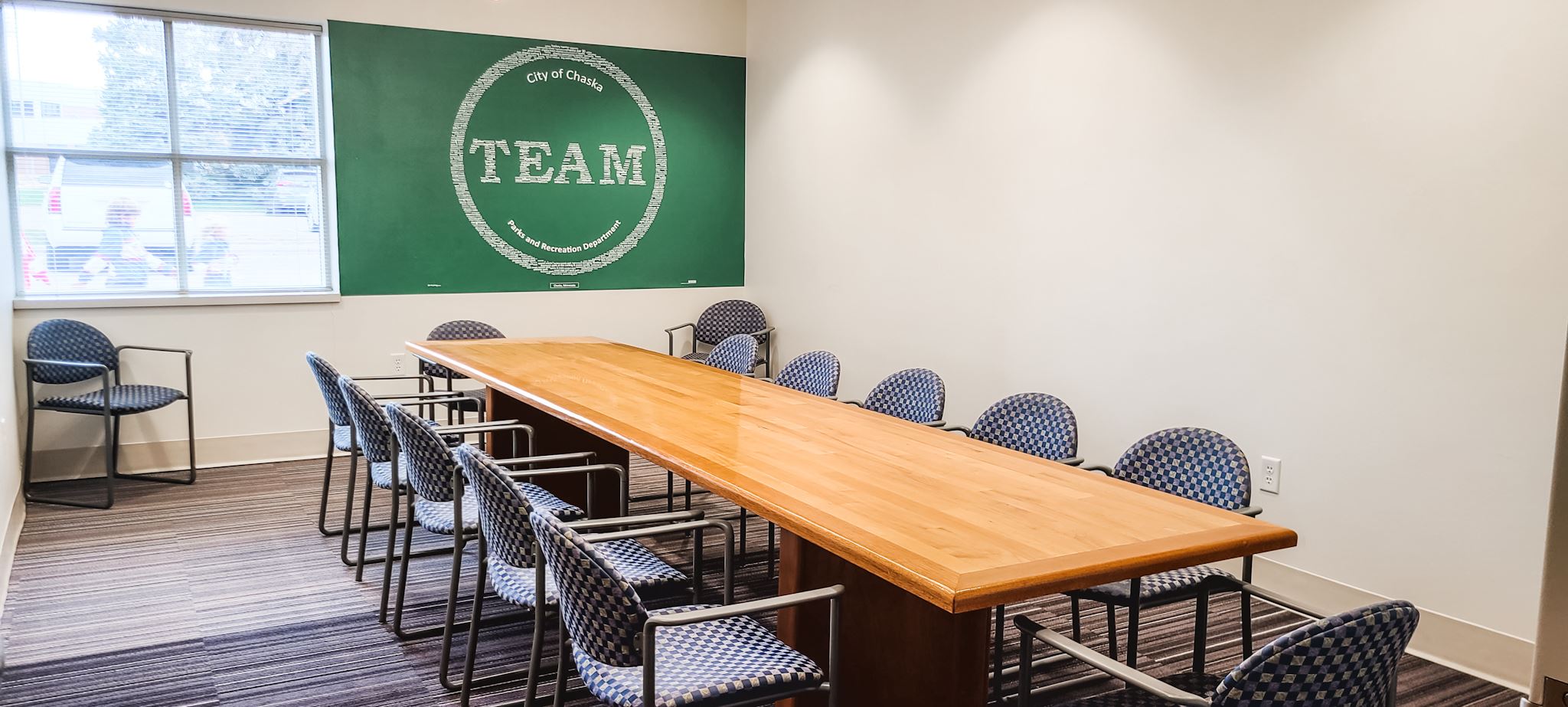 Photo depicts a wooden conference table with 12 chairs, the green Chaska logo and window on wall